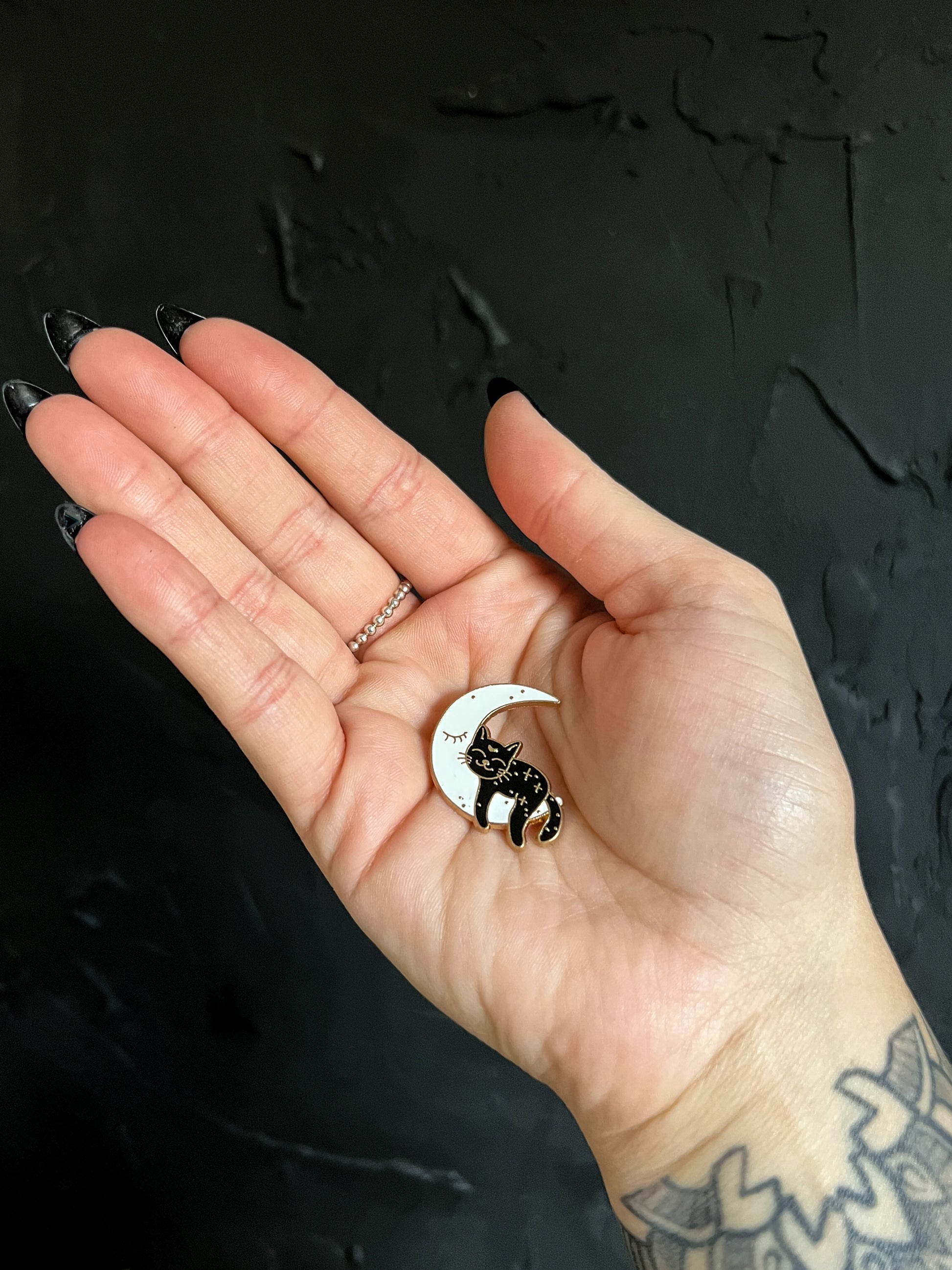 In the hand of The Stone Maidens, a really cute sleeping black cat on a white crescent moon enamel pin. 